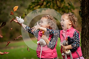 Identical twins having fun with autumn leaves, blond cute curly girls, happy family, beautiful girls in pink jackets