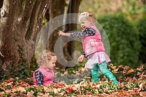 Identical twins having fun with autumn leaves in the park, blond cute curly girls, happy kids, beautiful girls in pink jackets