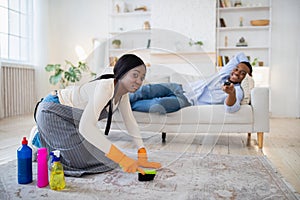 Idle black man watching TV, lying on sofa, while his exhausted unhappy wife cleaning home, copy space