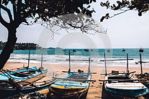 Idle fishing boats tied to the shore at a secluded beach in Sri Lanka. Blue ocean, windy weather and waves in the background.