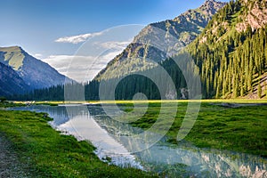 Idyllic summer landscape with hiking trail in the mountains with beautiful fresh green mountain pastures, river with reflection