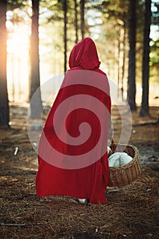 If we go out to the woods today...Rear view shot of a little girl in a red cape walking in the woods with a basket.