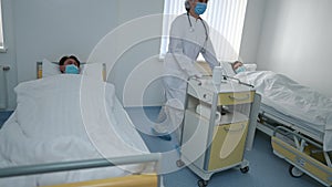 Ill Caucasian men in Covid-19 face masks lying in beds in hospital ward as nurse rolling bedside table leaving. Patients