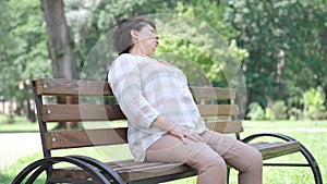 Ill Caucasian senior woman having heart attack sitting on bench in summer park as people passing by. Portrait of