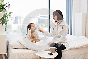 Ill girl resting on bed while doctor measuring temperature