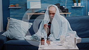 Ill man in blanket measuring temperature with thermometer