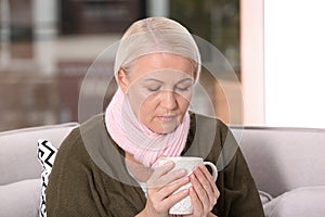 Ill mature woman with cup of hot tea for cough on sofa