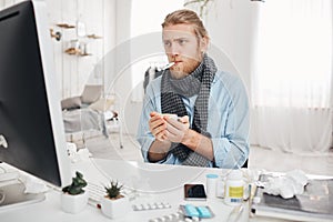 Ill sick bearded male sits in front of computer screen with thermometer in mouth, measures temperature, holds a cup of