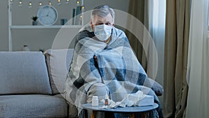 Ill sick old male in medical mask sitting on couch at home Caucasian elderly man wrapped in blanket feel unwell illness