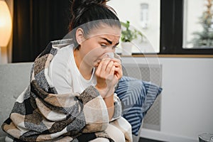 Ill young woman sit on sofa covered with blanket, freezing, blowing her runny nose, sneeze in tissue. Female got fever