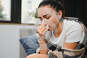 Ill young woman sit on sofa covered with blanket, freezing, blowing her runny nose, sneeze in tissue. Female got fever