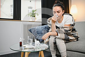 Ill young woman sit on sofa covered with blanket, freezing, blowing her runny nose, sneeze in tissue. Female got fever