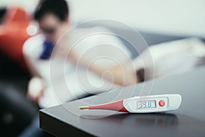 Illness and fever: Thermometer and ill man drinking tea in the blurry background