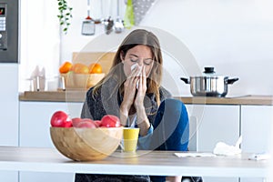 Illness young woman sneezing in a tissue while sitting in the kitchen at home