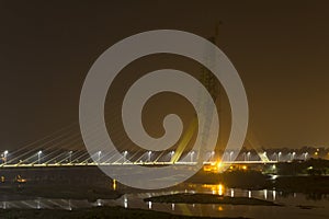 Illuminated cable-stayed bridge under construction with a tower crane over the Yamuna River at night. Signature Bridge. Delhi