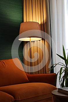 Illuminated floor lamp against beige curtains and green wall shining over orange sofa