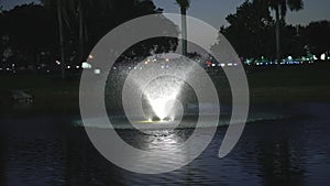 Illuminated fountain on the bay in an evening park with palm trees. There is a beautiful fountain in the evening park by