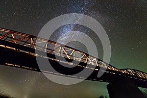 Illuminated metal bridge on concrete supports reflected in water on dark starry sky with Milky Way constellation background. Night
