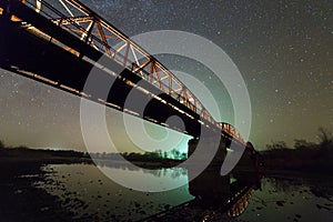 Illuminated metal bridge on concrete supports reflected in water on dark starry sky with Milky Way constellation background. Night