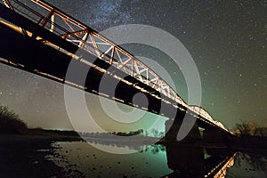 Illuminated metal bridge on concrete supports reflected in water on dark starry sky with Milky Way constellation background. Night