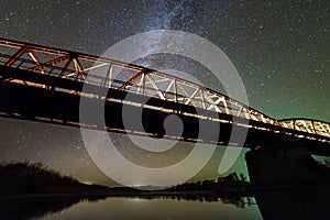 Illuminated metal bridge on concrete supports reflected in water on dark starry sky with Milky Way constellation background. Night