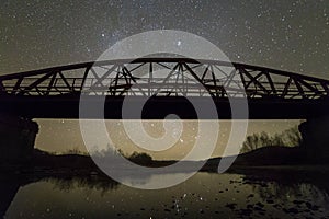 Illuminated metal bridge on concrete supports reflected in water on dark starry sky with Milky Way constellation background. Night