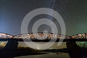 Illuminated metal bridge on concrete supports reflected in water on dark starry sky with Milky Way constellation background. Night