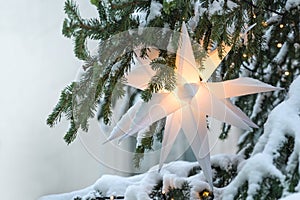 Illuminated white Moravian star (German Herrnhuter Stern) hanging in a snow covered Christmas fir tree