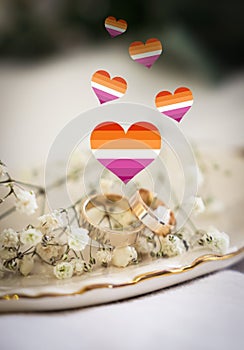 Illustration of hearts in color of lesbian flag, beautiful wedding rings and gypsophila flowers on white cloth, closeup view