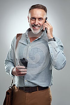 Im on my way. Studio portrait of a mature man talking on a cellphone while carrying a bag and cup of coffee against a