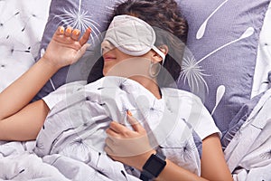 Image of adorable girl covering her eyes with sleeping mask and lying on pillow in bed, lady does not want to wake up early in