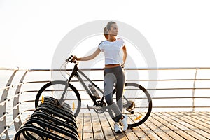 Image of adorable woman standing with bicycle on boardwalk, during sunrise over sea