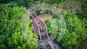 Aerial view above large metal bridge for train tracks over river surrounded by green trees