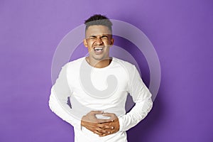 Image of african-american man in white sweatshirt, touching belly and screaming in pain, having stomach ache, food