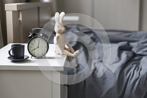 Image alarm clock with a black cup of coffee on white bedside table in front of the bed with gray linens. The room is in beige