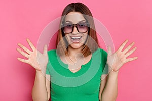 Image of amazed pleasantly surprised girl with brown hair wearing green casual t shirt and sunglasses posing against pink wall,