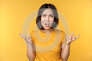 Image of angry asian woman, shouting and cursing, looking outraged, furious face expression, standing over yellow