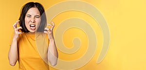 Image of angry asian woman, shouting and cursing, looking outraged, furious face expression, standing over yellow