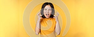 Image of angry asian woman, shouting and cursing, looking outraged, furious face expression, standing over yellow