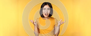 Image of angry asian woman, shouting and cursing, looking outraged, furious face expression, standing over yellow
