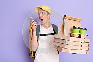 Image of angry emotional young adult man courier in yellow cap and T-shirt holding pizza boxes and coffee to go isolated on purple