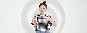 Image of angry young woman complains, points at telephone and looks disappointed, isolated against white background