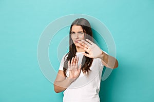 Image of annoyed brunette woman in white t-shirt, defending herself, raising hands to protect eyes from bright light