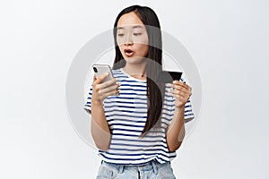 Image of asian girl holding credit card, looks excited at mobile phone screen, paying for online shopping, using store