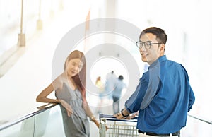 Image of asian mid aged shopper man leaning on shopping cart, moving on walkway and smiling, his girlfriend in the background