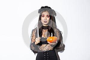 Image of asian woman panicking, being scared of witchcraft on halloween, standing in witch costume with candle and