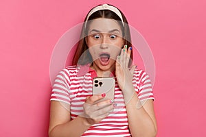 Image of astonished amazed young girl with brown hair wearing striped casual t shirt, using cell phone, reading message with