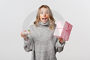Image of attractive blond girl celebrating birthday, looking surprised, holding b-day cake and a gift, standing over