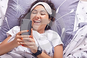 Image of attractive brunette girl lying on pillow with smart phone in hands, wearing t shirt and blindfold, female smiling happily