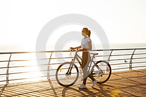 Image of attractive woman standing with bicycle on boardwalk, during sunrise over sea